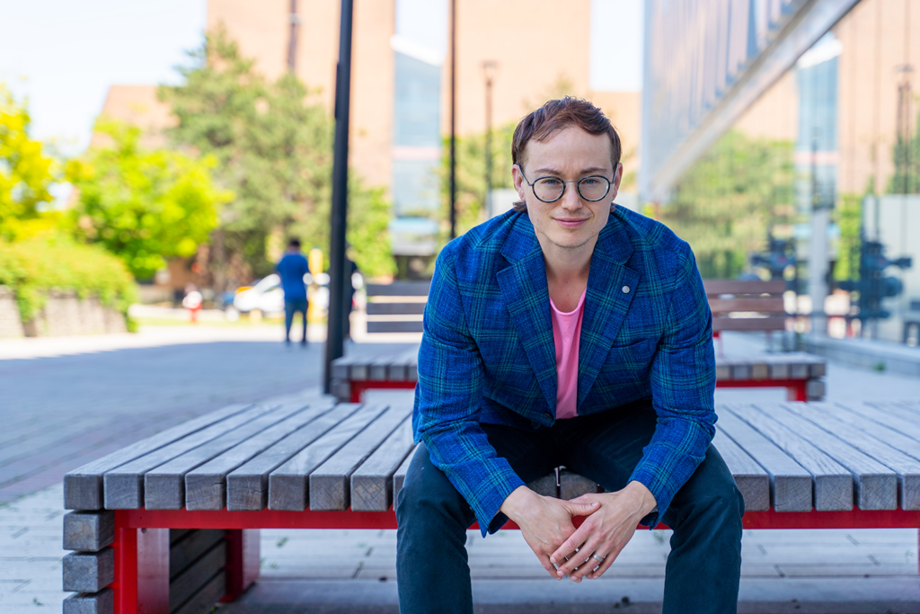 An image of Professor Kinnon MacKinnon sitting on a bench, leaning forward. Professor MacKinnon is wearing a blue plaid coat with a pink t-shirt underneath. 