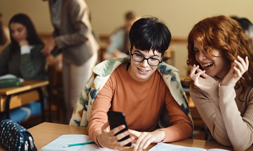 Happy female high school students reading a text message on smart phone during a class in the classroom.