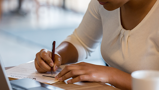 An unrecognizable Hispanic woman references the information on her laptop to fill out a job application at the coffee shop.