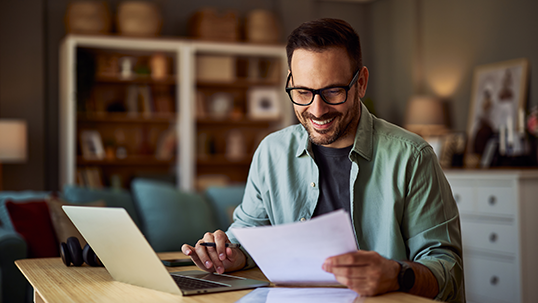 A smiling male college professor reading a student's essay paper in his office in front of a laptop
