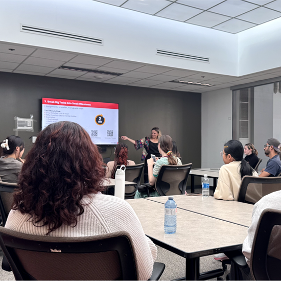 Participants seated around tables listen to a presenter pointing to a slide displayed on a screen during a workshop session.