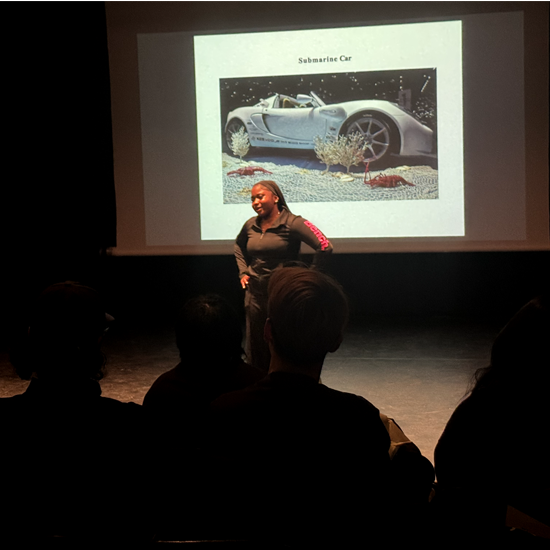 A presenter stands on a stage in front of a projected slide while audience members watch from their seats during an academic event.