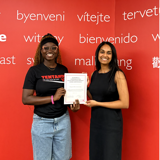 Two individuals stand in front of a red wall displaying greetings in multiple languages while holding a certificate of achievement.