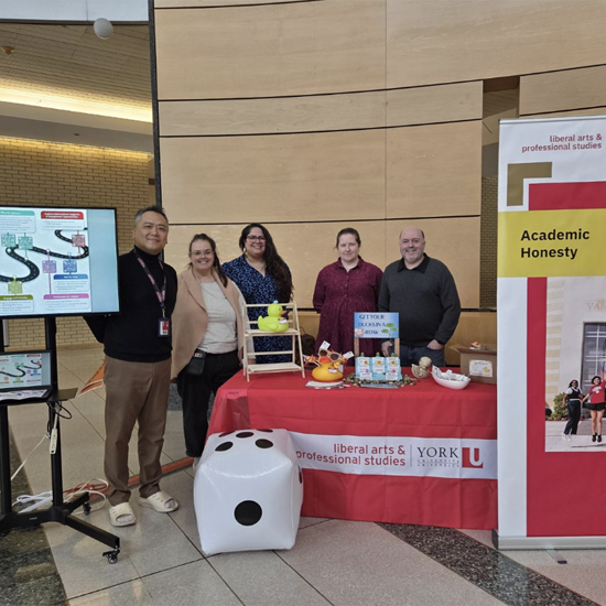 Staff members pose behind an Academic Honesty information table featuring educational resources, giveaways, and display materials in a campus atrium.