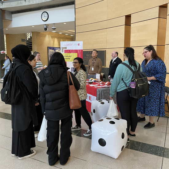 Students gather around an Academic Honesty information table in a campus atrium, speaking with staff members and viewing educational materials. Large inflatable dice are displayed beside the table.