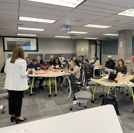 A facilitator stands at the front of a classroom leading a workshop while participants seated at round tables listen, take notes, and work on activities.