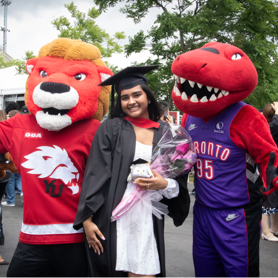 A graduate wearing a gown and cap poses for a photo between two university mascots while holding a bouquet of flowers and a small gift.
