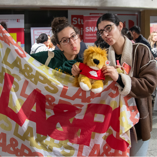Two attendees pose with a stuffed lion mascot while holding a large LA&PS banner at a campus event, with information booths visible in the background.