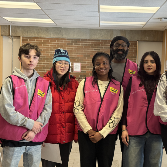 Five volunteers wearing pink event vests pose for a group photo indoors during a First Year Lunch Break program