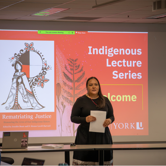 A speaker stands in front of a presentation slide reading “Indigenous Lecture Series Welcome” while addressing attendees in a classroom setting.