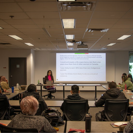 Attendees seated around tables listen to a presentation during an Indigenous Lecture Series event, with presentation slides projected on a screen at the front of the room.