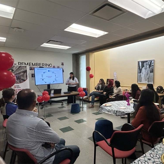 Attendees seated in a library event space listen to a presentation, with a speaker standing beside a screen displaying presentation slides.