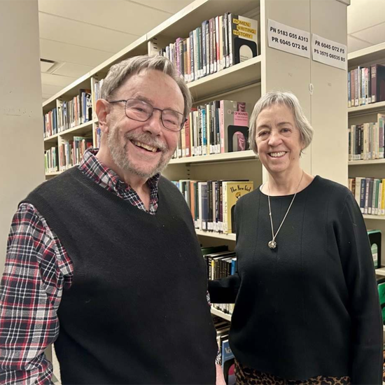 Two individuals stand among library bookshelves posing for a photo in a library setting.