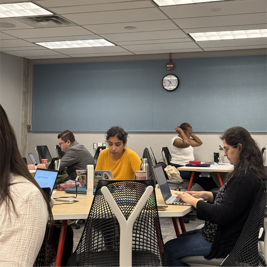 A group of participants gathered around a table engage in discussion while working on laptops and reviewing printed materials during a collaborative workshop session