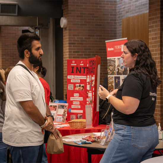 Two people speak at an information booth displaying resources and promotional materials during an orientation event held in an indoor common area.