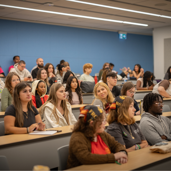 Attendees seated in a large lecture hall listen to presenters standing at the front of the room, with a projection screen displaying orientation information.