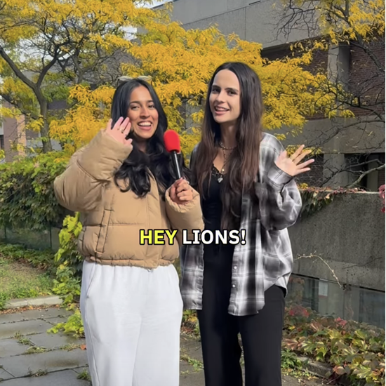 Two people standing outdoors among yellow autumn trees, waving toward the camera while one holds a microphone, with the text “HEY LIONS!” on screen.