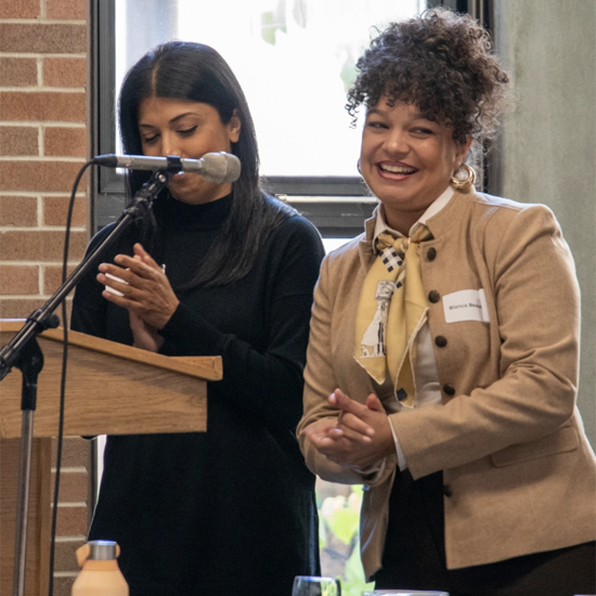Two speakers stand at a podium during an academic event, with one person speaking into a microphone while the other stands nearby in front of a large window.