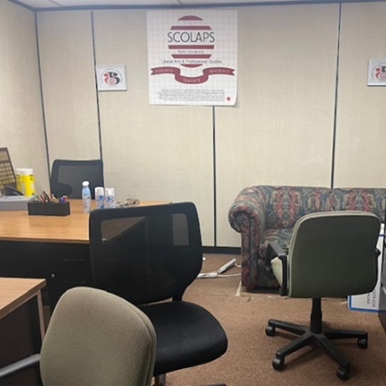 A student lounge and office space before renovation, featuring office chairs, desks, a patterned couch, and a SCOLAPS sign mounted on the wall.