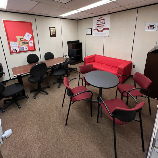 A renovated student lounge featuring red seating, round tables, meeting chairs, and a SCOLAPS sign on the wall, arranged for student gatherings and study sessions.