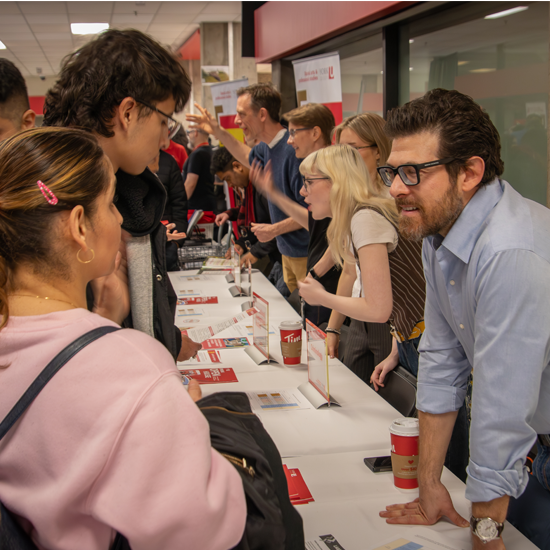 Prospective students and university representatives speak across an information table covered with brochures and program materials during a campus open house event.