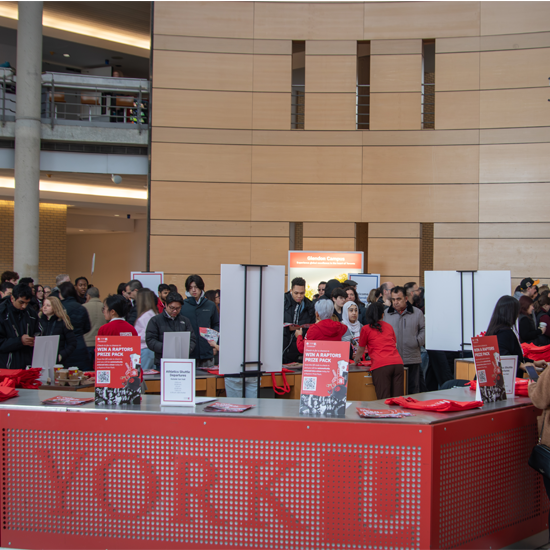 Visitors gather in a large campus atrium around registration and information stations during a busy open house event.
