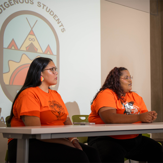 Two presenters wearing matching orange shirts sit at a table during a discussion session, with an Indigenous Students Association logo projected on a screen behind them.