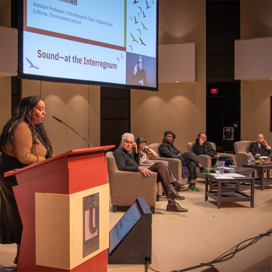 A speaker stands at a podium addressing an audience while a panel of speakers sits on stage beneath a projected lecture presentation.