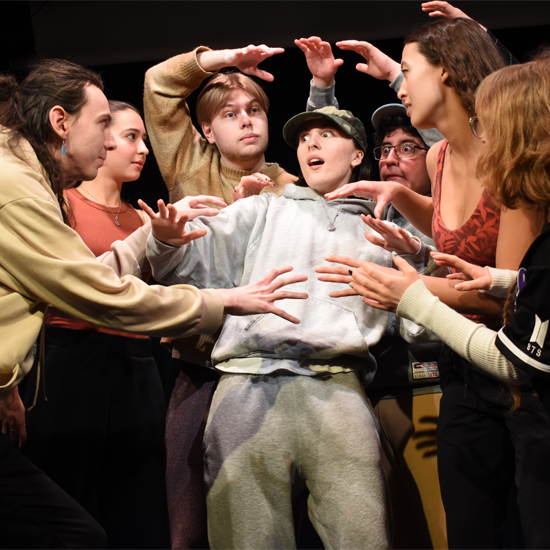 Participants on a theatre stage perform a dramatic scene, with one person at the center surrounded by others extending their arms as part of a group performance.