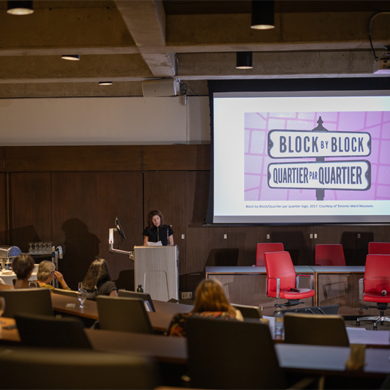 A presenter stands at a podium addressing attendees in a lecture hall while a large screen displays a slide about the Block by Block project.