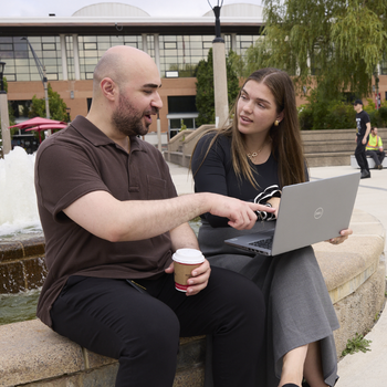 Two people discuss a project while reviewing a laptop beside a campus fountain
