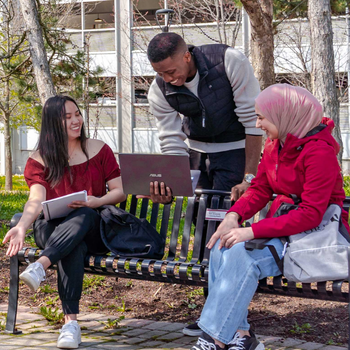 Students gather outdoors on campus, collaborating around a laptop and notebooks
