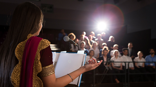 Young woman stands at podium to read a presentation in front of an audience