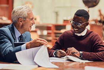 Older man helps young student work on writing assignment in while sitting in a library