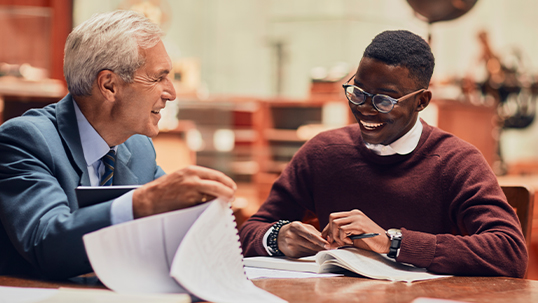 Older man helps young student work on writing assignment in while sitting in a library