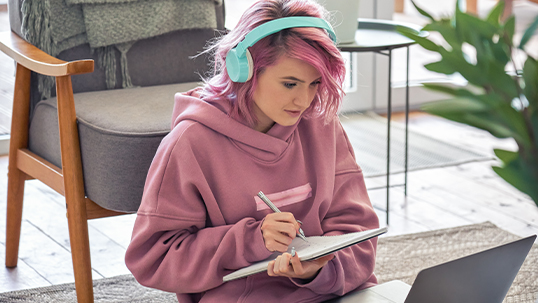 image of a young woman wearing headphones while she holds a pen and notebook and looks at her laptop screen sitting on a rug