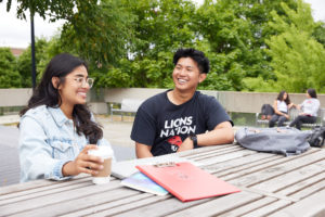 Girl and boy smiling at each other while sitting outside at a picnic table. There are notebooks on the table.