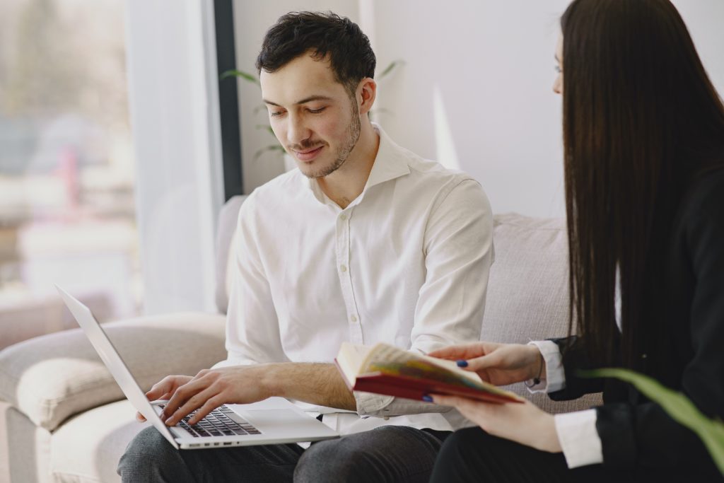 Man and woman sitting on a couch. The man is typing on his laptop while the woman looks at him with a book in her hands