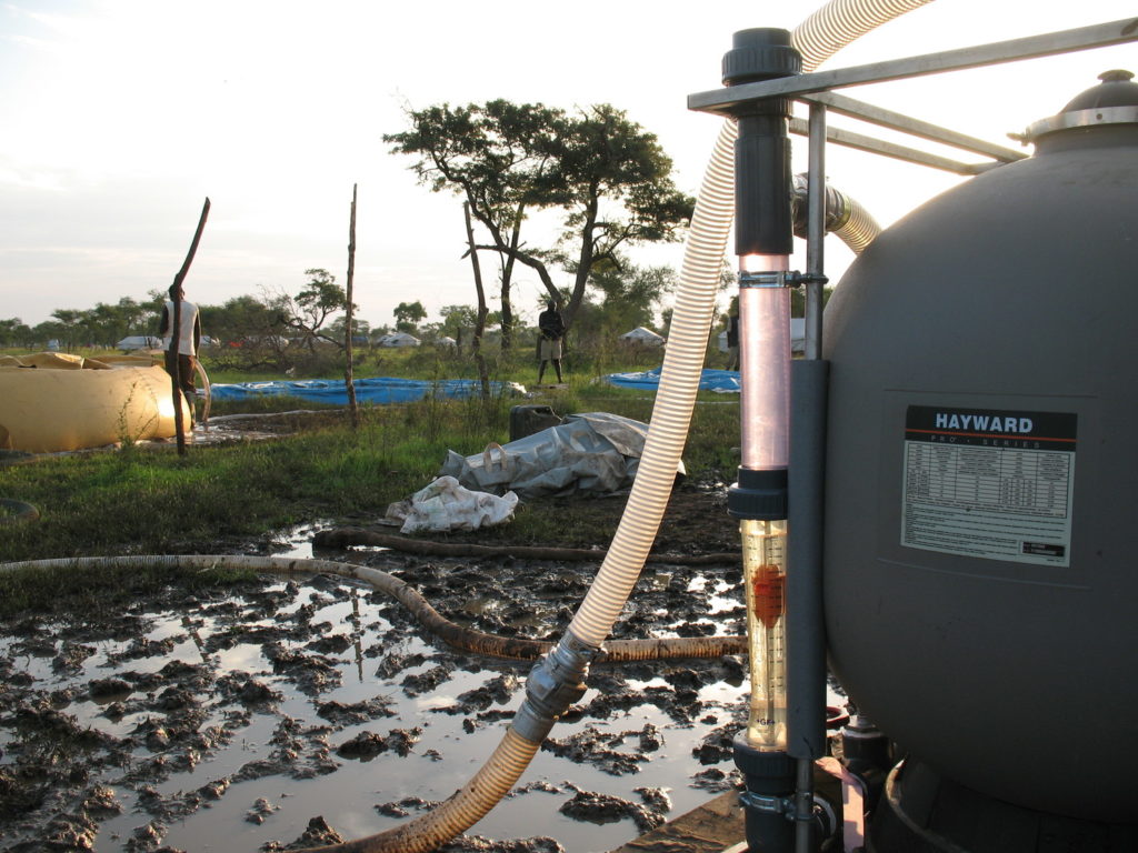 A water filter that says "Hayward" on it is seen in the foreground on flooded rural land with trees and men standing in the background.