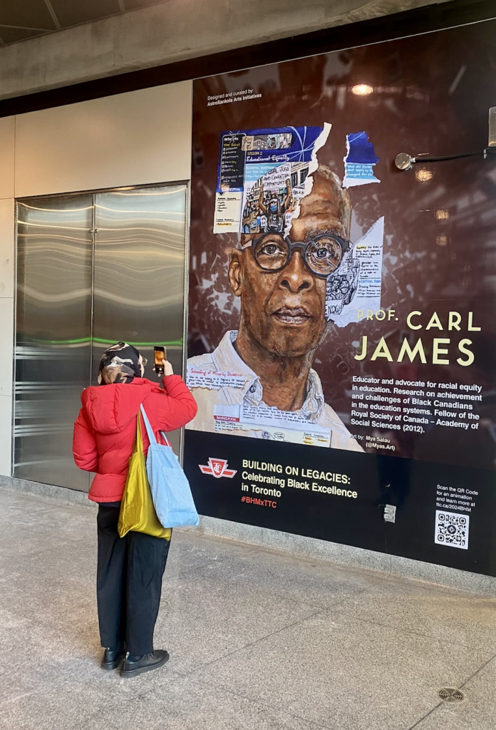 A person takes a photo of an art mural depicting Carl James at York University subway station.