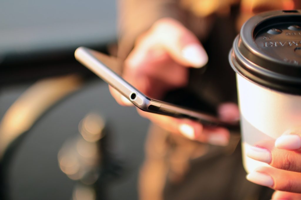 A close-up of a hand holding a cellular phone and the other holding a cup of coffee.