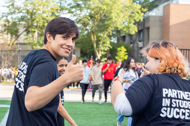 Two students with their backs to the camera, one of them is turning to look at the camera and give a thumbs up while smiling.