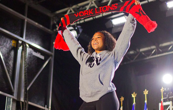 A student on stage at an Orientation event wearing a grey York University hoodie and holding read York University scarf above her head.