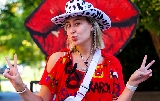 A girl in a red shirt with purse and cowboy hat flashes double peace signs at the camera.