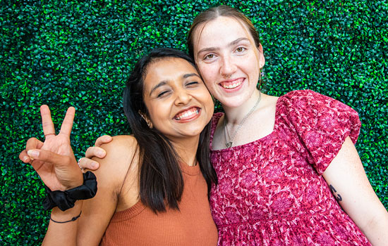 Two girls hug and smile in front of a green vegetation backdrop.