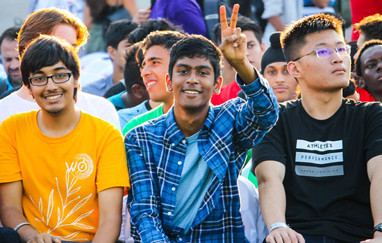 A group of students seated in the sun at an orientation event, one throwing up a peace sign to the camera.