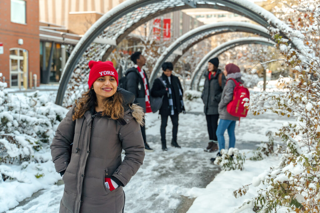 A girl wearing a red York toque stands outside on a snowy path while other students congregate and chat behind her.