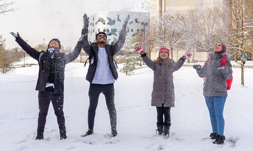 Four students stand in the middle of a snowy field and throw snow into the air.