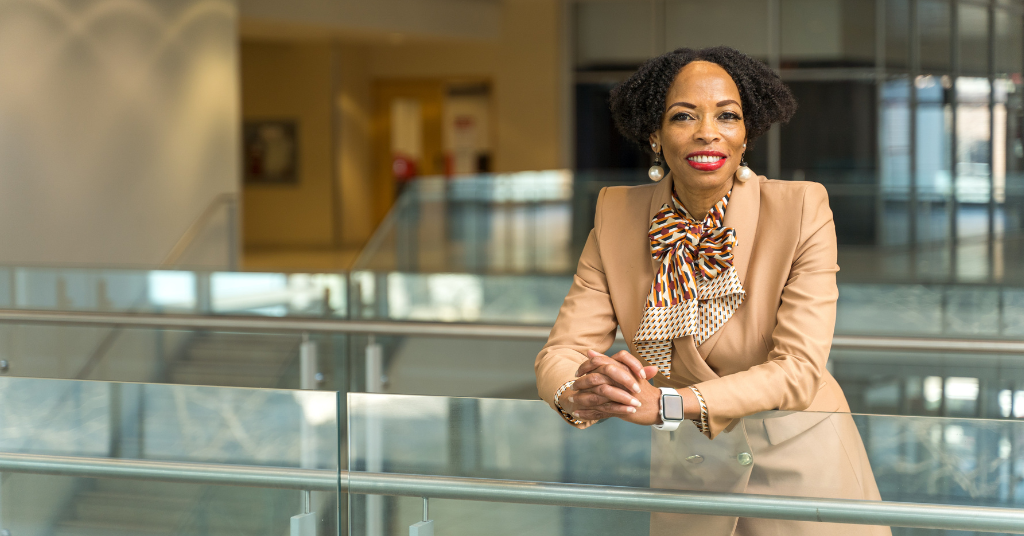 Andrea Davis, a Black female professor at York University, is leaning over a glass railing while wearing a beige colour suit, beige and brown printed silk scarf around her neck, red lipstick and large pearl drop earrings.