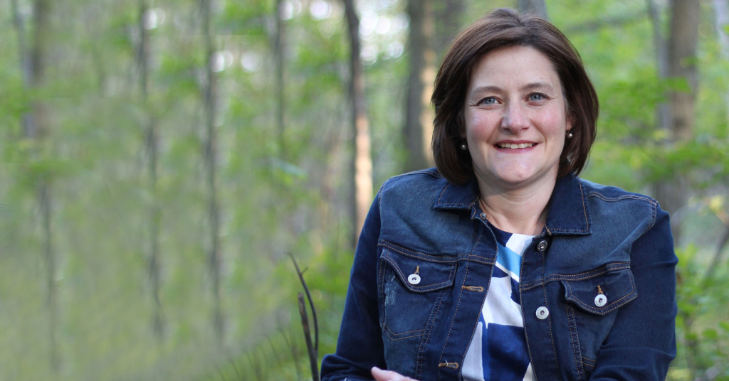 Nicole Arsenault learning towards the camera while wearing a blue and white top with a denim jacket among a forest with greenery in the background